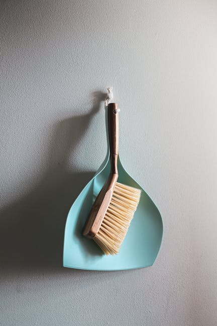A close-up of a light grey wall featuring a minimalist hanging arrangement consisting of a small blue plastic tray and a wooden scrub brush with bristles, both suspended by a single hook. The wall sur