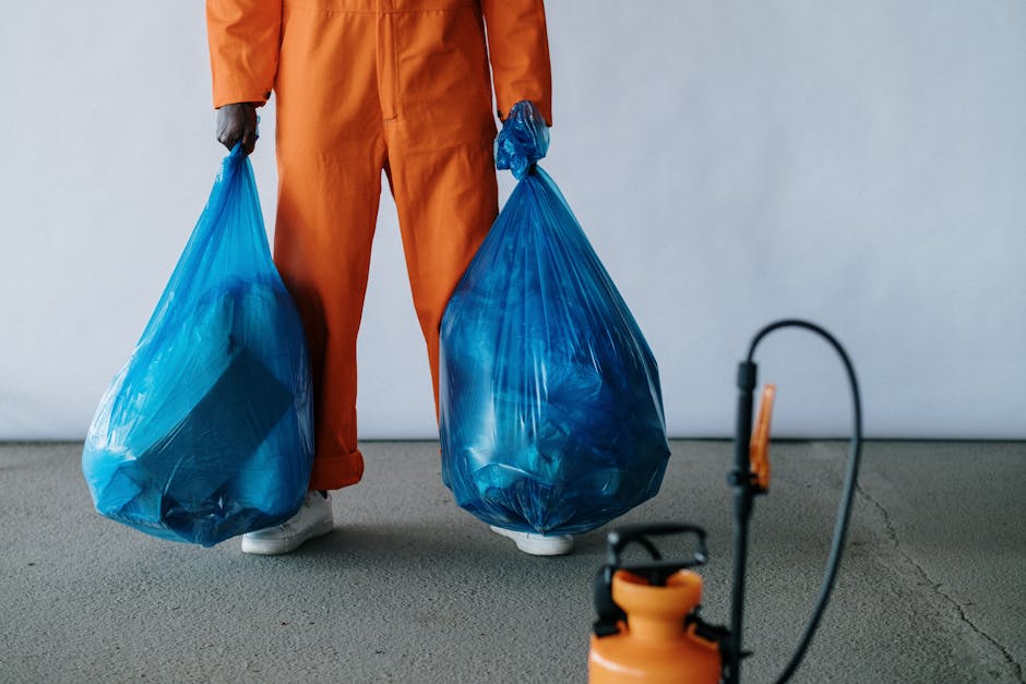 A person dressed in orange workwear standing on a grey concrete floor holding two large blue plastic garbage bags filled with waste, with their feet visible near the bottom of the frame. The backgroun