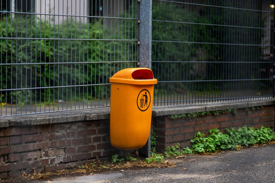 A person dressed in orange workwear standing on a grey concrete floor holding two large blue plastic garbage bags filled with waste, with their feet visible near the bottom of the frame. The backgroun