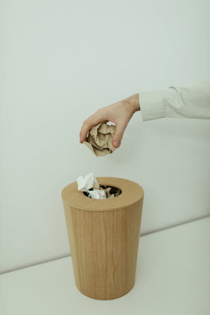 A person's hand, clad in a white long-sleeve shirt, is seen disposing of crumpled paper into a light wood, cylindrical waste bin placed against a plain white wall. The bin features a rounded top openi